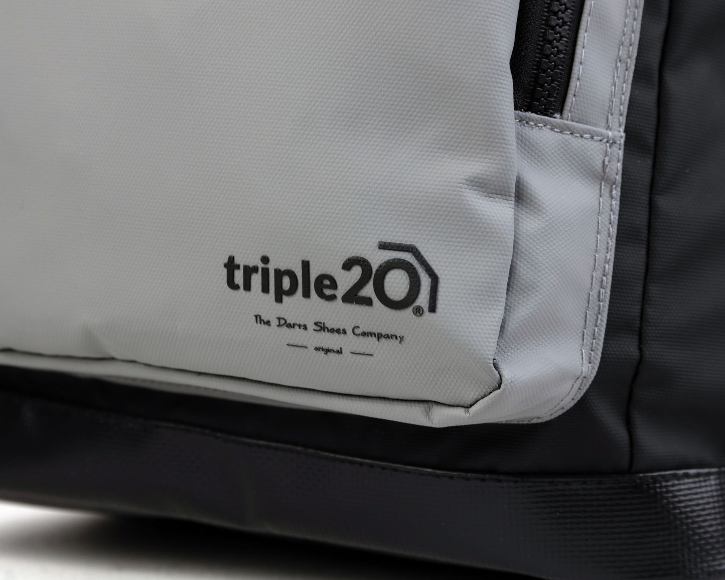 Hier ist die Nahaufnahme eines triple20 Dartrucksacks, welche die graue Vordertasche und das dort aufgebrachte Logo zeigt. Der dargestellte Rucksack wurde von einem Hersteller für Dartschuhe als Ergänzung zu den offerierten Dartschuhen in Deutschland designt. Der Hauptzweck ist die Aufbewahrung der Schuhe, natürlich lassen sich mit dem Rucksack auch Gegenstände des täglichen Bedarfs wie z.B. Einkäufe transportieren.