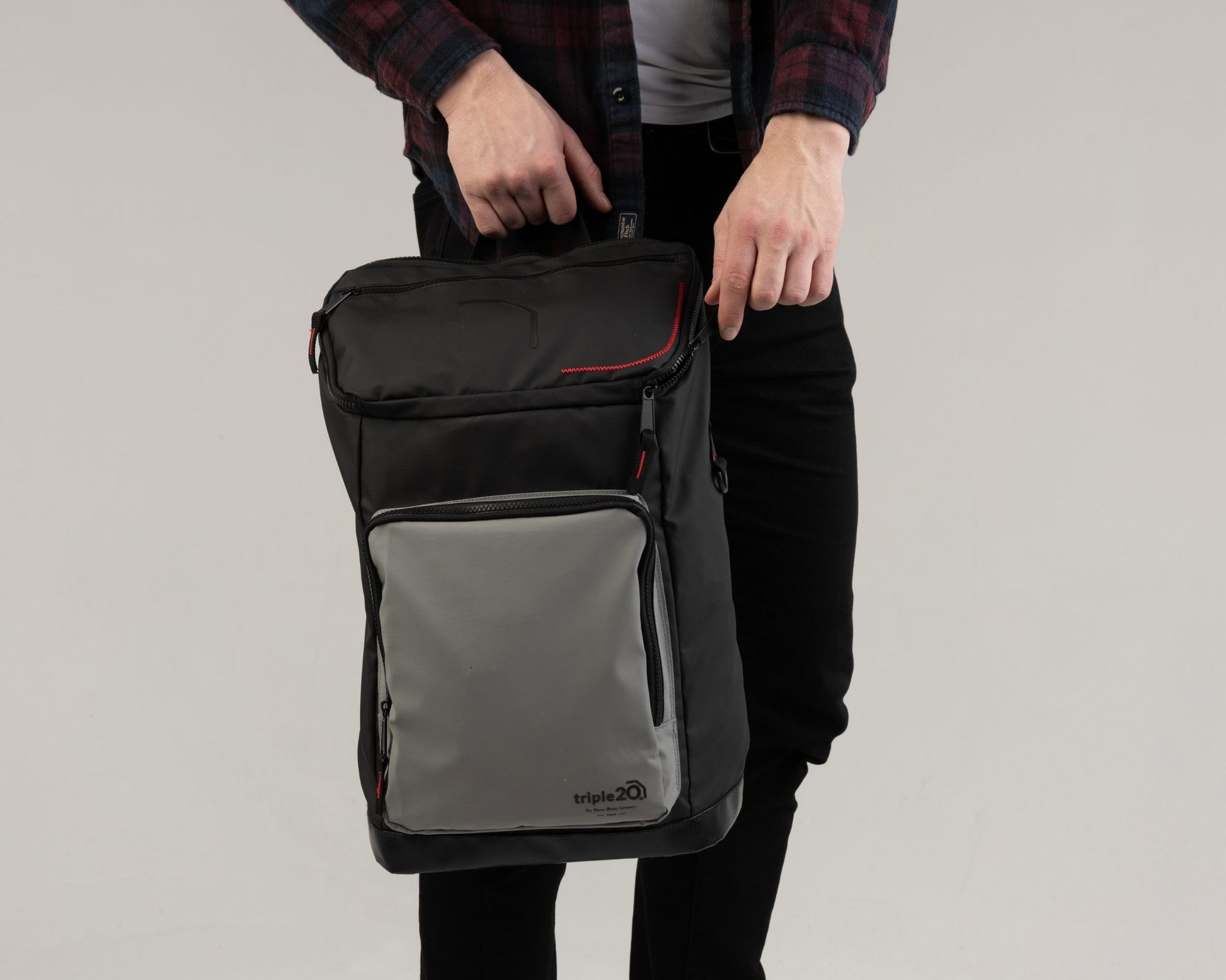 Produktfoto eines Dartrucksacks, der auf dem Foto von einem männlichen Model in den Händen gehalten wird. Es wird mit den Händen angedeutet wie der obere Reißverschluss geöffnet wird. Der Rucksack ist von der Grundfarbe her schwarz und hat eine hellgraue Fronttasche mit dem Logo des Herstellers, triple20. Als Highlight verfügt das Rucksackmodell über knallrote Details.